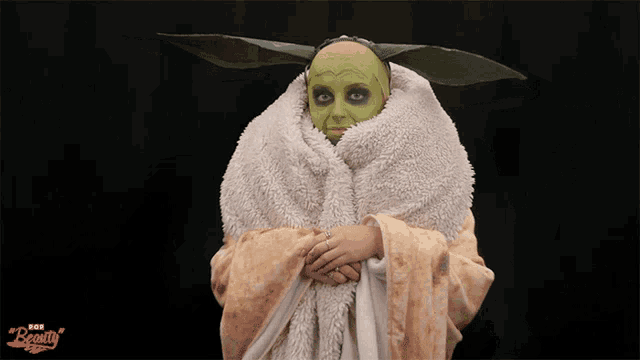 a woman is wrapped in a blanket and has a green face painted like baby yoda