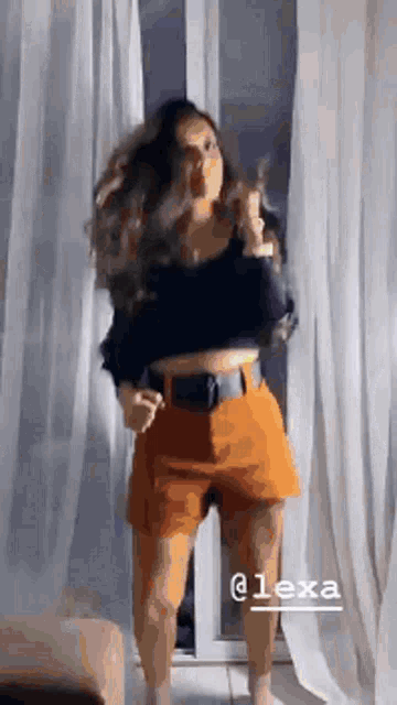 a woman in a black crop top and orange shorts is standing in front of a window .