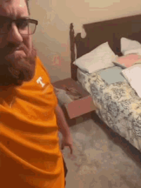a man with a beard and glasses is standing in front of a bed in a bedroom .