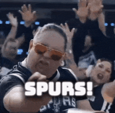 a man wearing sunglasses and a spurs shirt is standing in front of a crowd of people .
