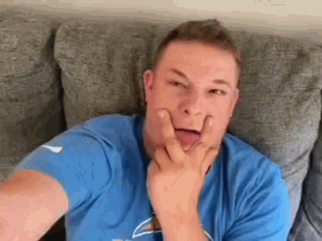 a man in a blue shirt is sitting on a couch making a funny face with his hands on his face .