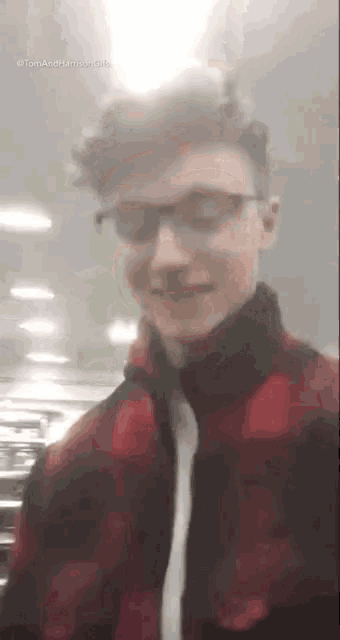 a young man wearing glasses and a red plaid jacket is standing in a room .