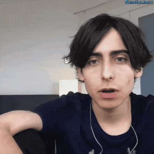 a young man wearing a blue shirt and ear buds looks at the camera with a sunstar.hero watermark on the bottom
