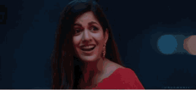 a woman in a red saree is smiling and looking at the camera in a dark room .