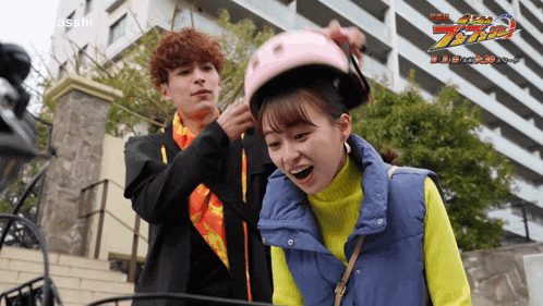 a man helps a woman put on a pink helmet with the word asahi on the bottom right
