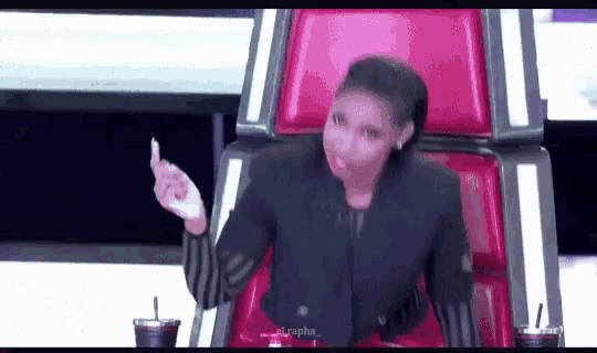 a woman in a black jacket is sitting in a red chair and giving a middle finger .