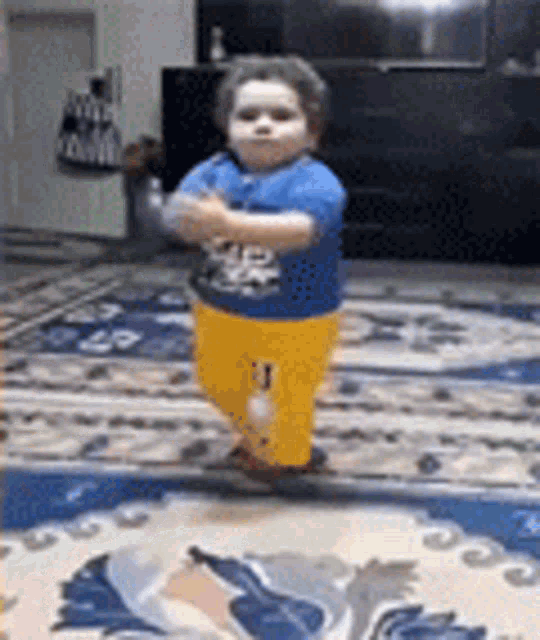 a baby in a blue shirt and yellow pants is standing on a rug in a living room .