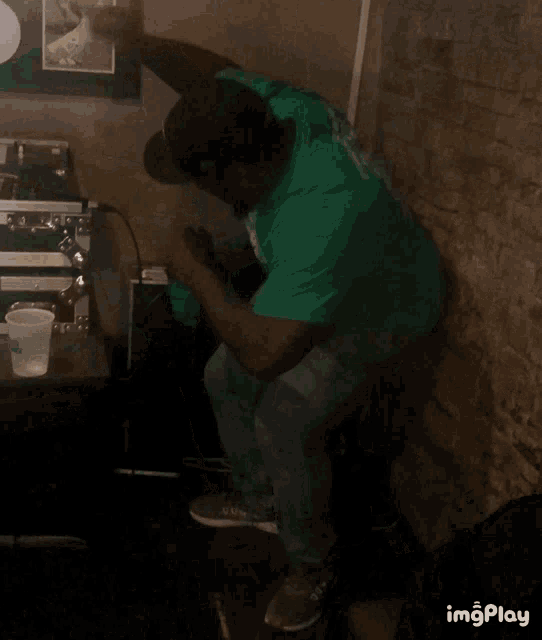 a man in a green shirt is sitting in front of a brick wall with imgplay written on the bottom right