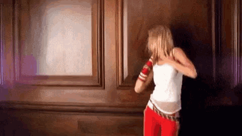 a woman in a white tank top and red pants is dancing in a room in front of a wooden wall .