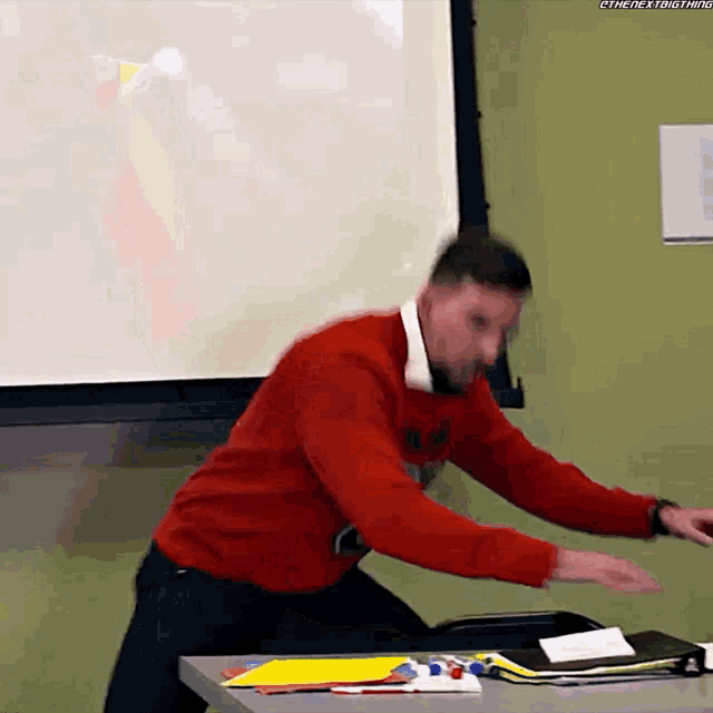 a man in a red sweater is standing in front of a screen that says " the next thing " on it
