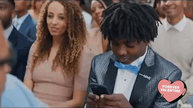 a man in a tuxedo and bow tie is looking at his cell phone while sitting in a crowd of people .