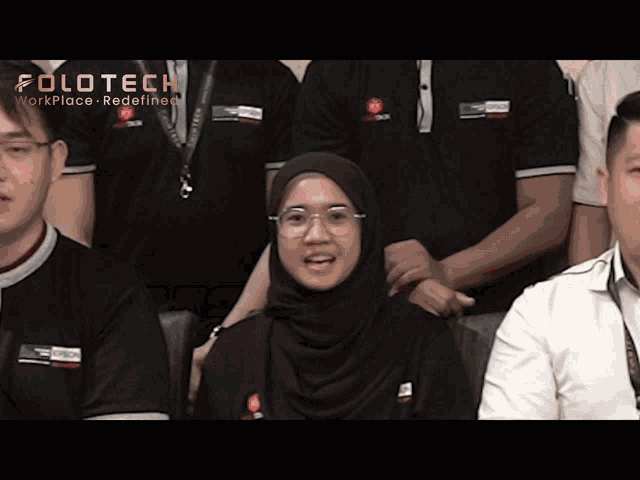a woman wearing a hijab and glasses stands in front of a group of polotech employees