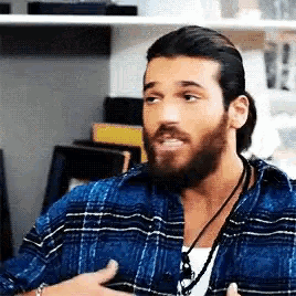 a man with a beard wearing a plaid shirt and a necklace is sitting in a room .