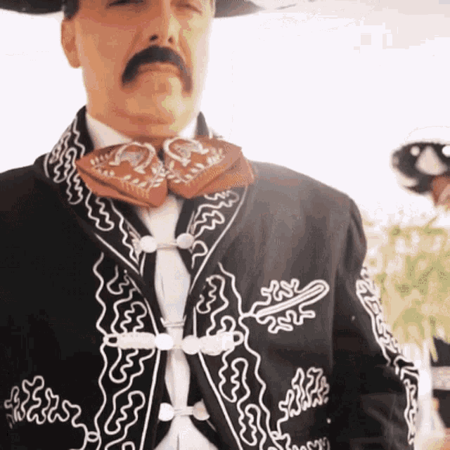 a man with a mustache is wearing a black jacket with white embroidery