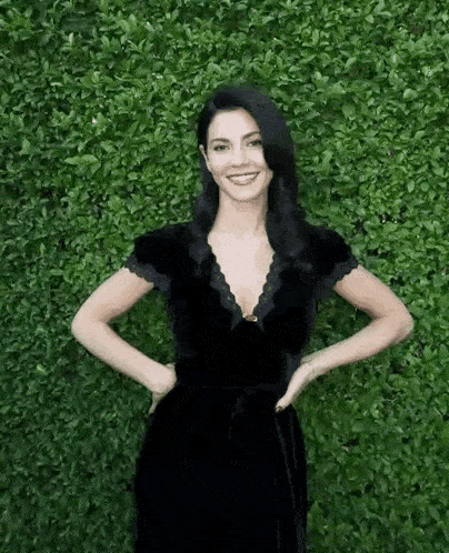 a woman in a black dress is posing for a picture in front of a green hedge .
