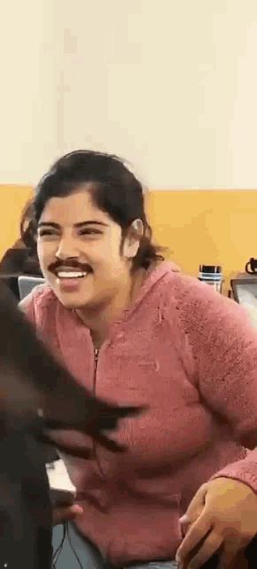 a woman wearing a pink sweater and a mustache is smiling and laughing .