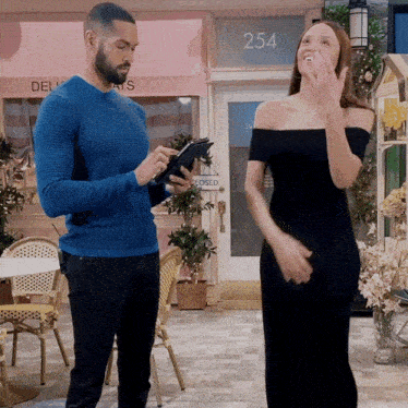 a man in a blue sweater holds a tablet in front of a woman in a black dress
