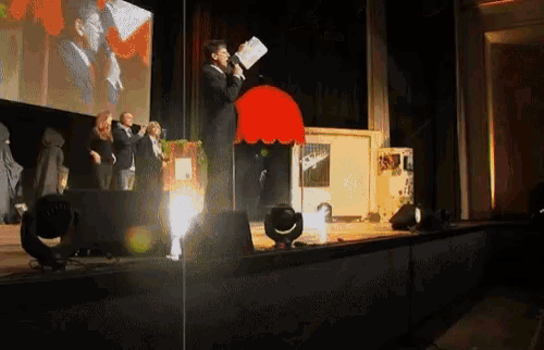 a man stands on a stage holding a piece of paper in front of a red lamp