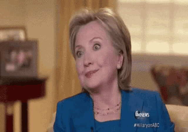 hillary clinton is sitting on a couch in a living room and smiling .
