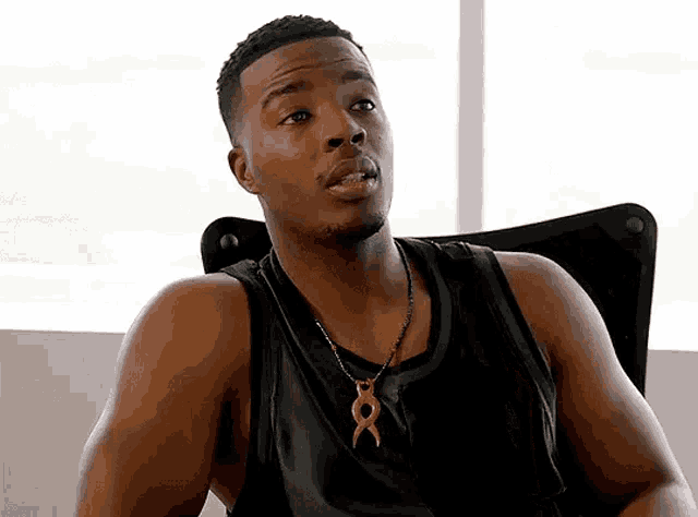 a man wearing a black tank top and a necklace with an x on it is sitting in front of a window