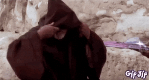 a man in a hooded robe is standing in front of a rock wall holding a sword .