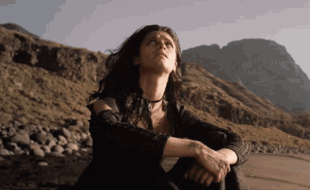 a woman sitting on a rocky beach looking up at the sky