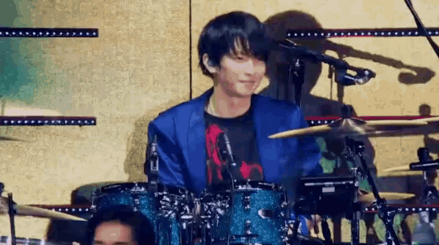 a man in a blue jacket is playing drums on a stage in front of a microphone .