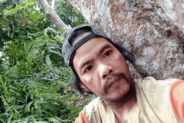a man with a beard wearing a baseball cap and a towel leaning against a tree