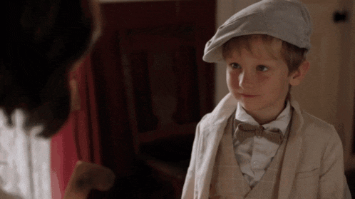 a young boy wearing a hat and bow tie looks at a woman