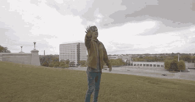 a man in a brown jacket is standing on top of a grass covered hill waving his hand .