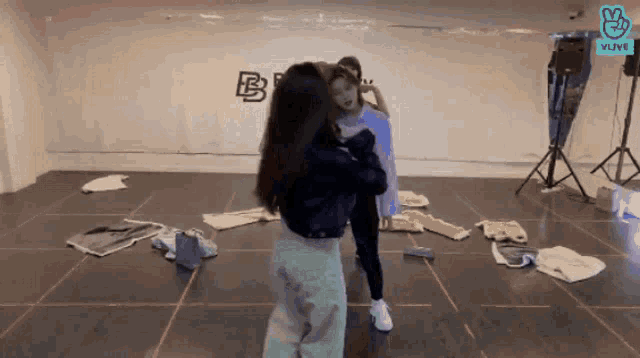 two women are dancing in a room with clothes on the floor and a vlive logo on the wall