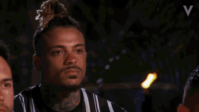 a man with dreadlocks and a tattoo on his face looks at the camera with the letter v above him