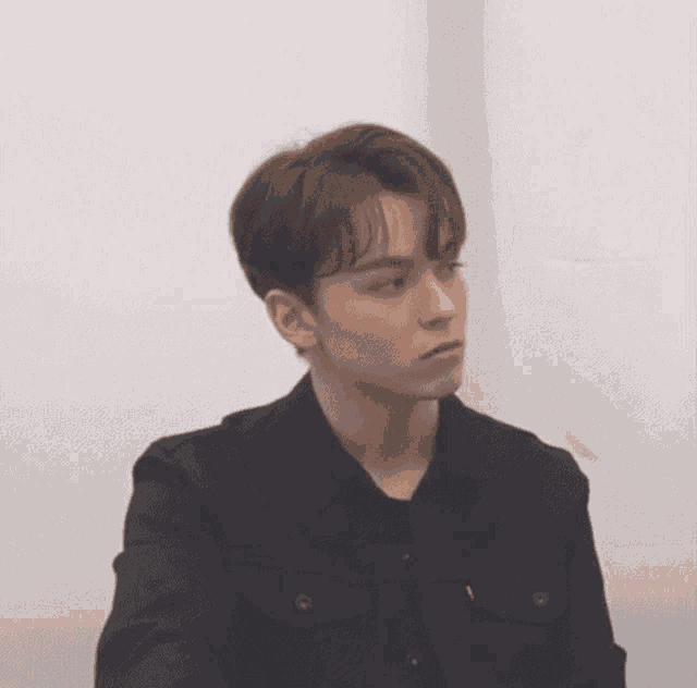 a young man wearing a black jacket and a black shirt is sitting in front of a wall .