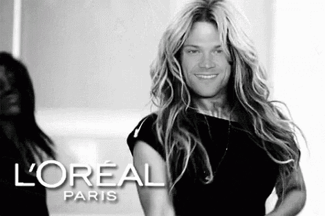 a man with long blonde hair is wearing a black dress and smiling .