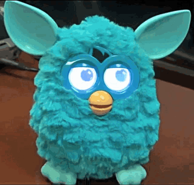 a blue furby with a yellow beak is sitting on a wooden table