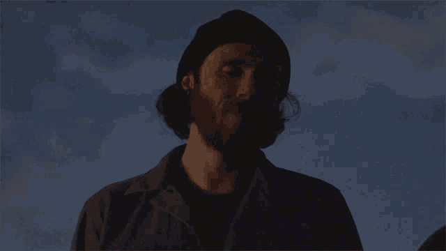 a man with a beard wearing a black hat stands in the dark