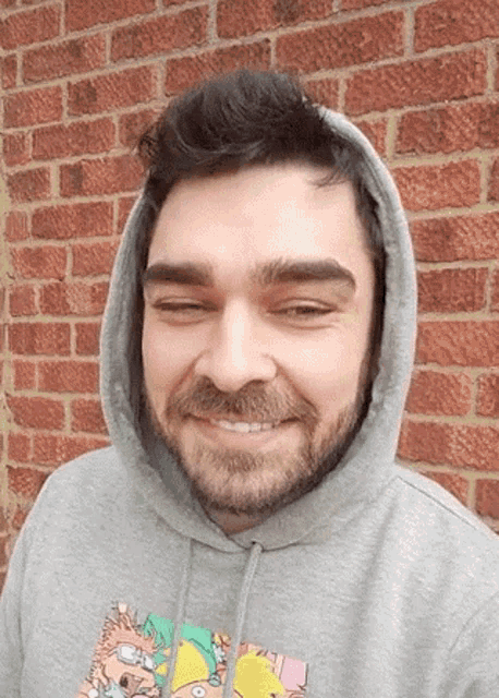 a man with a beard wearing a grey hoodie is smiling in front of a red brick wall