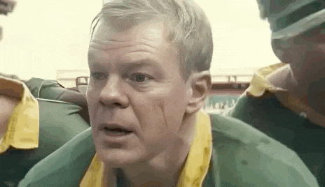 a man in a green shirt and yellow collar is standing in a crowd of people and looking at the camera .