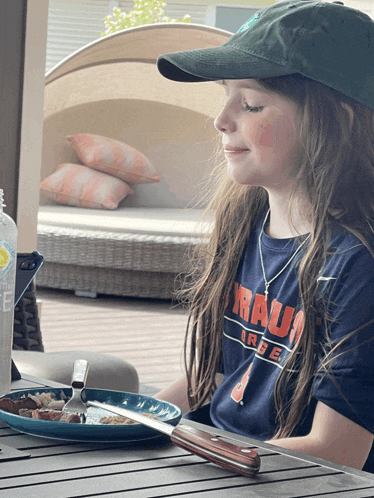 a girl wearing a blue shirt that says rau on it sits at a table with a plate of food