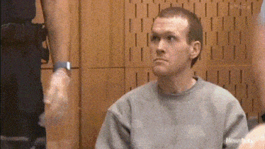 a man in a gray sweater is sitting in a courtroom with a police officer in the background .