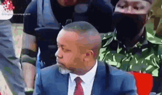a man in a suit and tie is sitting in front of a crowd of people wearing masks .