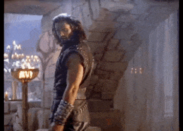 a man with long hair and a beard is standing in a stone room with a torch .