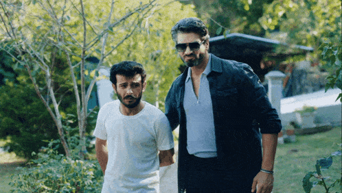 two men standing next to each other one wearing sunglasses and a black jacket