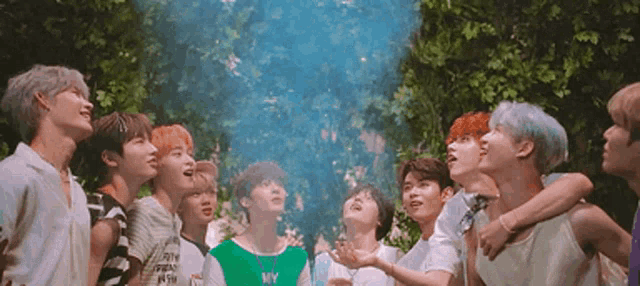 a group of young men are standing in a forest looking up at a blue smoke coming out of a tree .