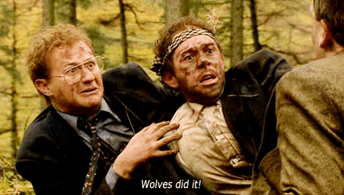 a man in a suit and tie holds another man in a forest and says " wolves did it "
