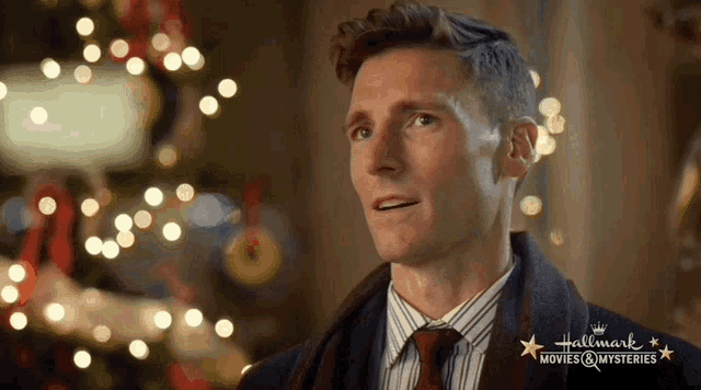 a man in a suit and tie is standing in front of a christmas tree and a sign that says hallmark movies & mysteries