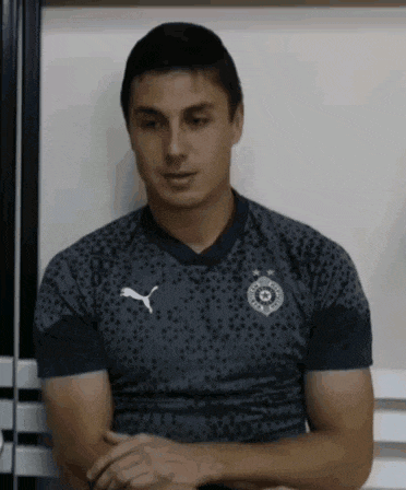 a man wearing a gray shirt with a puma logo on it is sitting with his arms crossed .