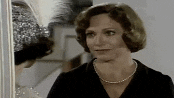 a woman in a black dress and pearl necklace is standing in front of a mirror .