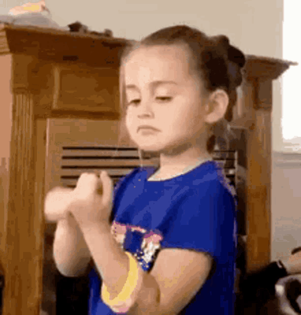 a little girl wearing a blue shirt and a yellow bracelet is standing in front of a fireplace in a living room .