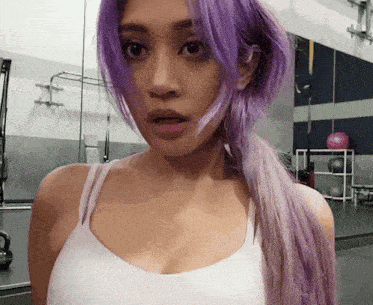 a woman with purple hair and a white tank top looks at the camera